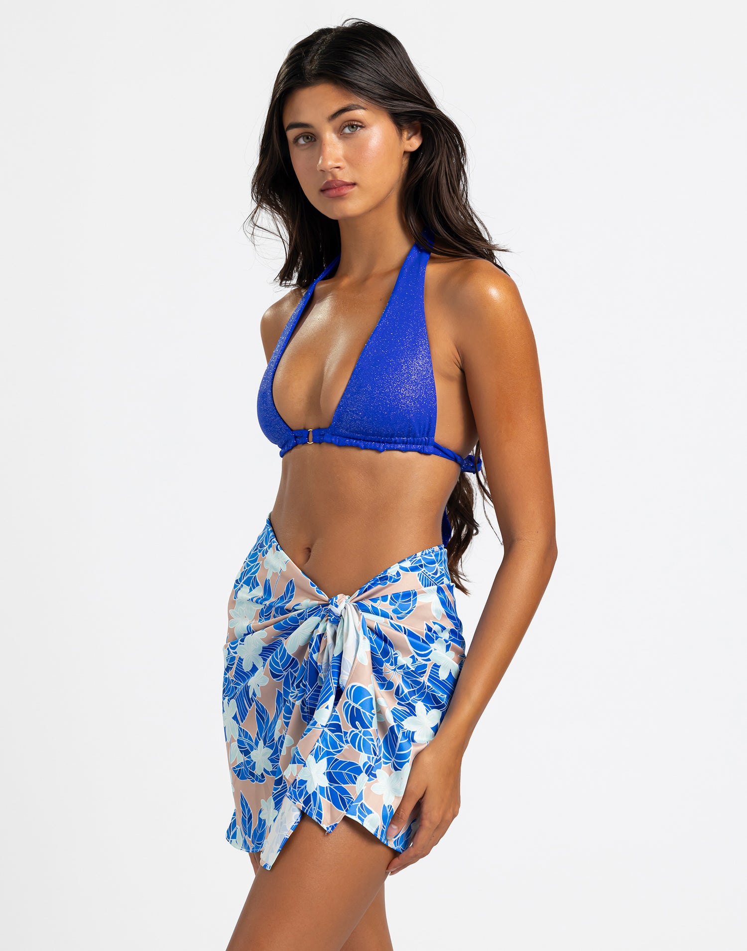 The model is wearing a short pareo with a vibrant blue floral print on a light background, tied at the waist for an effortlessly chic and beach-ready look.